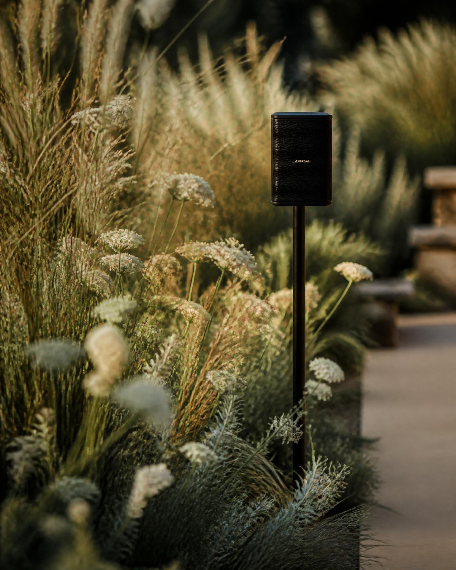 A compact black speaker on a slim stand tucked behind ornamental grasses and white florals at an outdoor ceremony